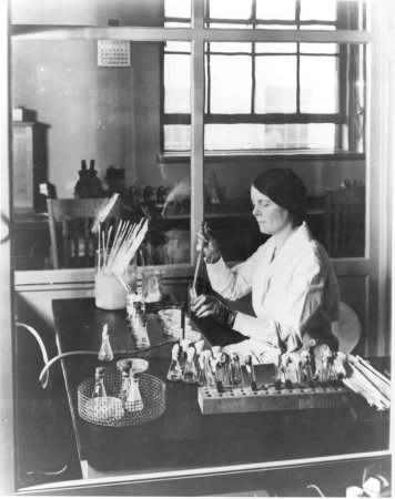 Black and white image of a woman sitting at a table performing experiments
