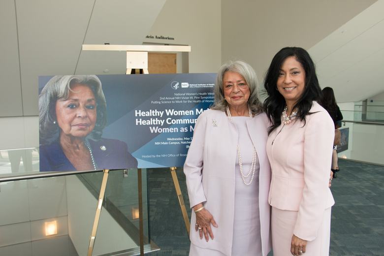 Two women pose in front of a poster. The poster has an image of the older woman and identifies the event as the Vivian Pinn Symposium. Both women are wearing light pink skirt suits.