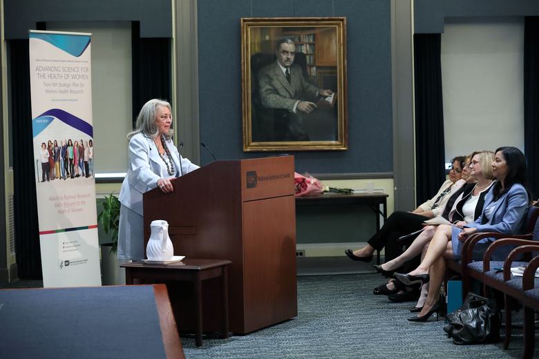 A woman stands behind a podium to the left of the image. In front of her is a row of seats. 