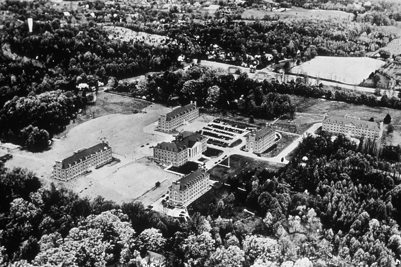 Black and white aerial image of 6 buildings and the roads and woods surrounding them