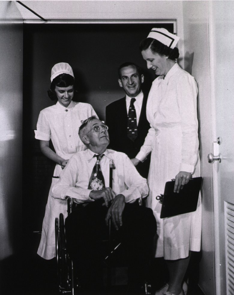 Black and white photo of a man in a wheel chair, two nurses, and a man in a suit