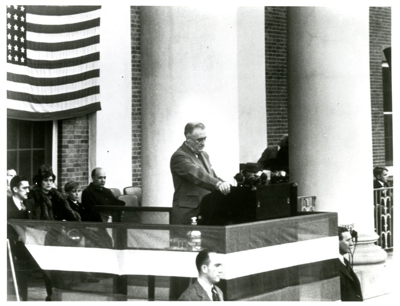 Black and white image of a man in a suite standing at a podium with an American flag behind him