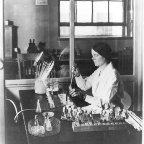Black and white image of a woman sitting at a table performing experiments